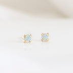 A pair of stud earrings with an Australian opal in a gold setting, placed on a white surface.