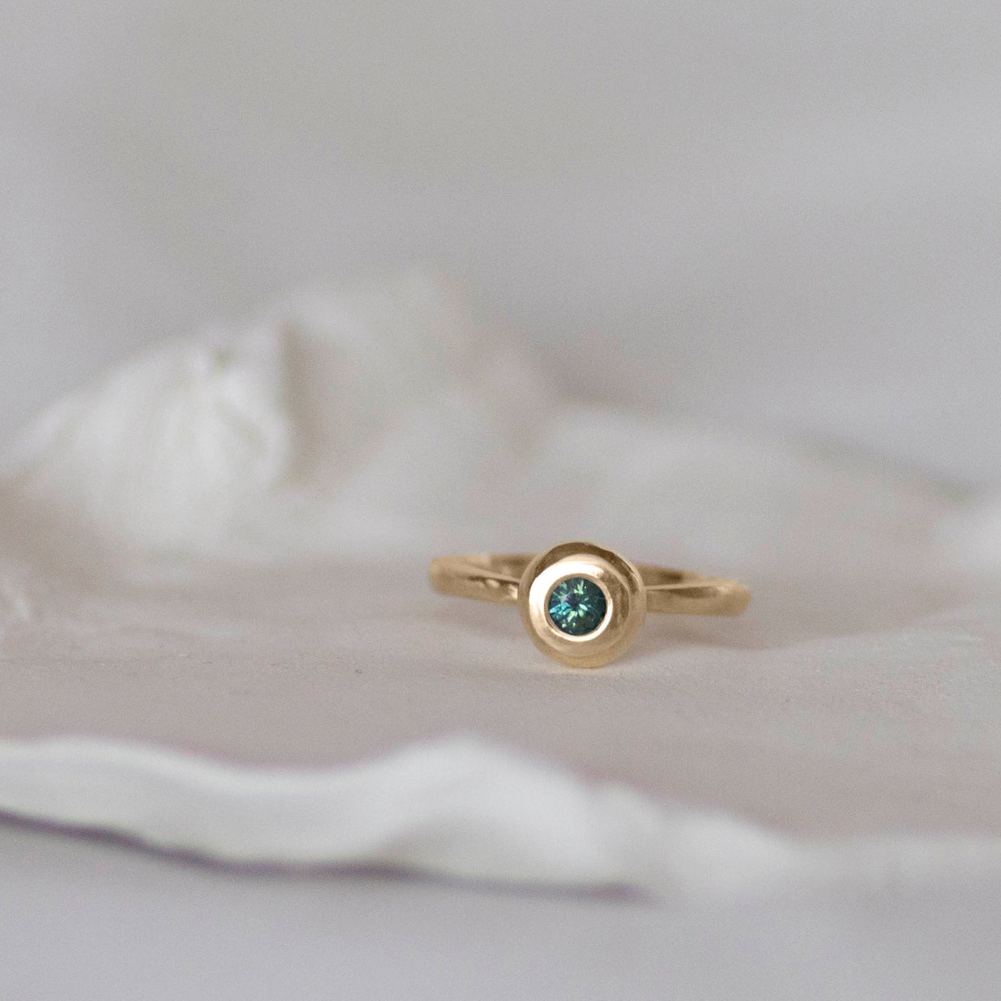 Australian Blue/Green Parti Sapphire set in a heavy, 14ct yellow gold ring sitting on a white ceramic dish by jane finch